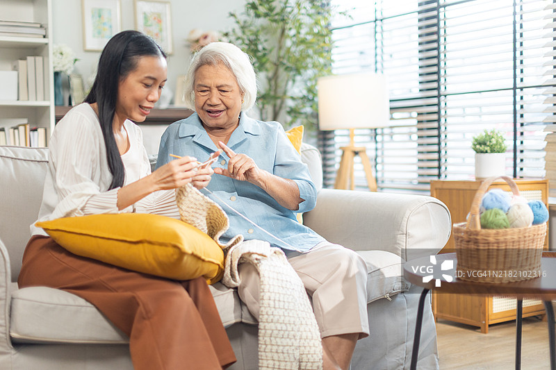 白发的亚洲老母亲在温馨的客厅里为她成年的女儿钩织围巾。母亲指导女儿编织。图片素材