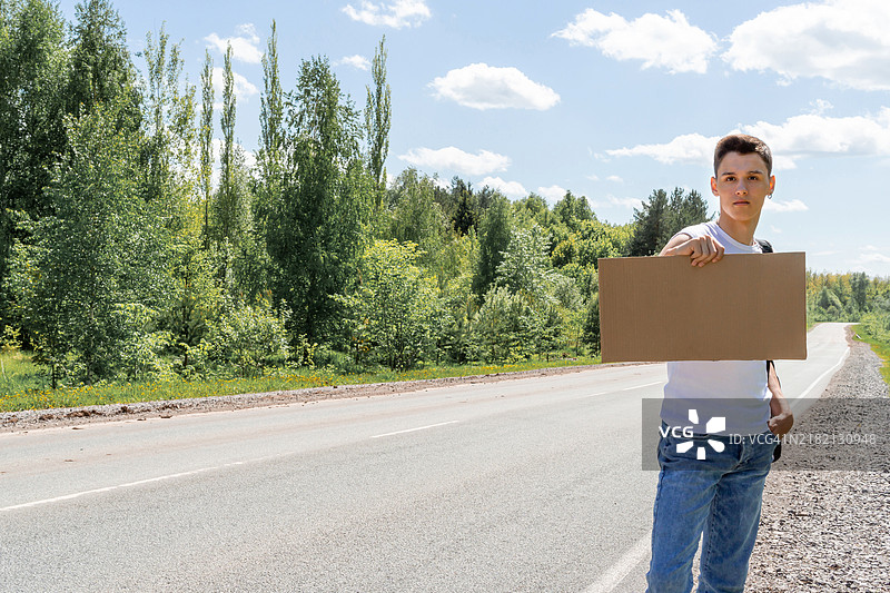 一个年轻人手持纸板标志在搭便车，标志上留有文字空间，表达对旅行的热情。图片素材