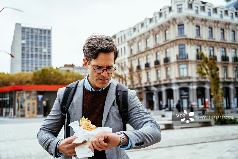 一位手拿三明治的商人在城市风景中走过，戴着眼镜，穿着商务休闲服，背景是办公大楼。图片素材