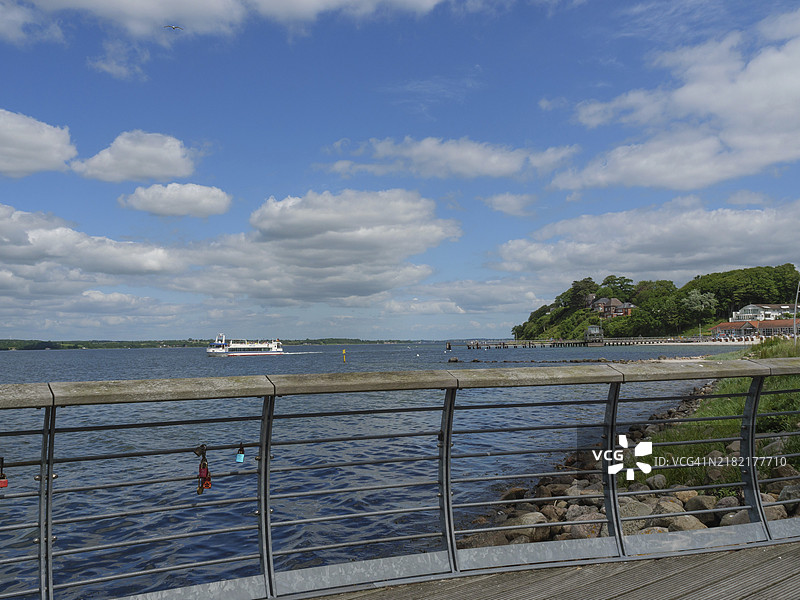 一条沿海的有栏杆的小路,远处有一艘船和树木茂密的海岸线,格吕克斯堡,石勒苏益格-荷尔斯泰因,德国图片素材