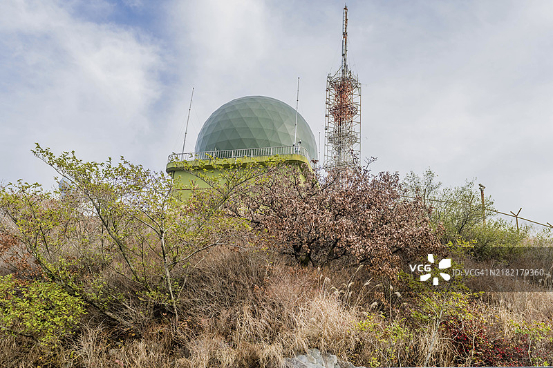 韩国一座长满杂草的山丘上，雷达站与绿色圆顶和通信塔并排而立，背景是多云的天空。图片素材