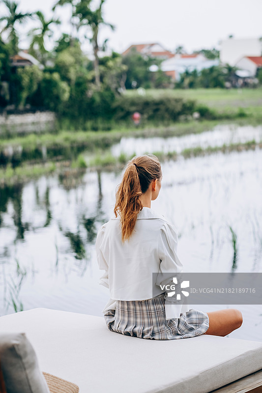 宁静湖边的平静反思：在大自然中放松和正念。一个扎着马尾的女人坐在白色长椅上，面朝湖水，姿势放松。图片素材