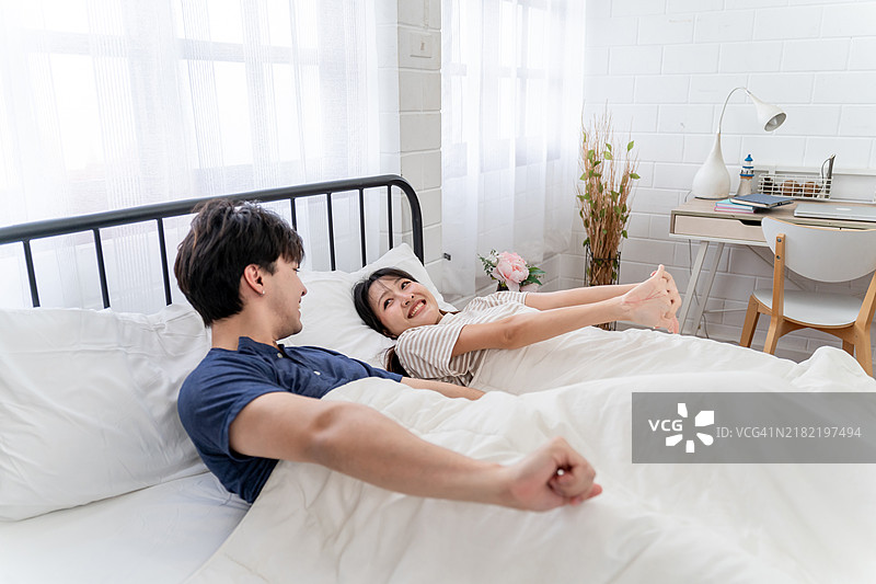 亚洲年轻情侣在家中床上醒来后躺着。迷人的男女感到快乐和放松，享受彼此交谈，度过早晨的闲暇时光。图片素材