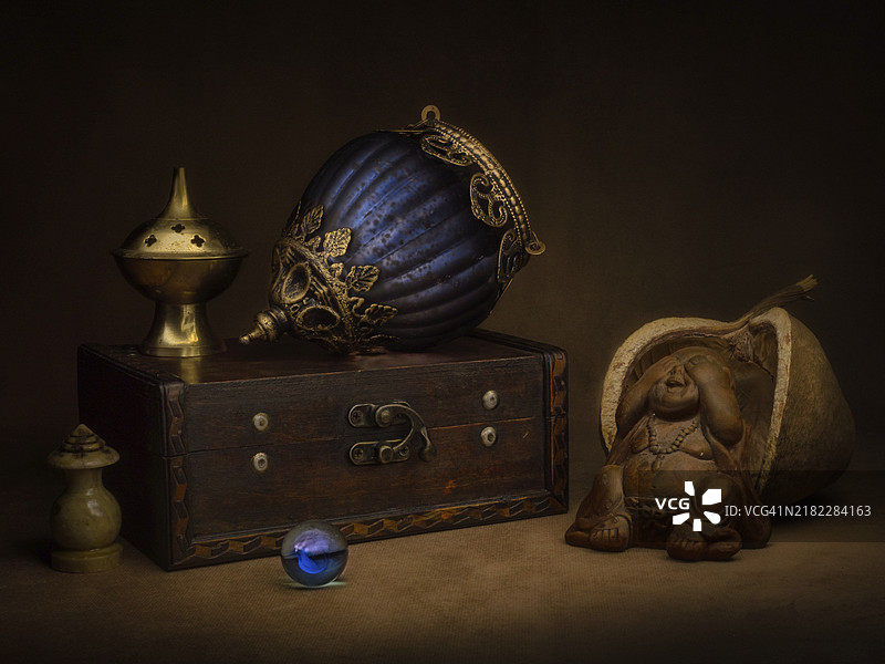 静物作品，包含香炉和东方吊灯放在木箱上，前面有玻璃球和笑佛，背景为深色。图片素材