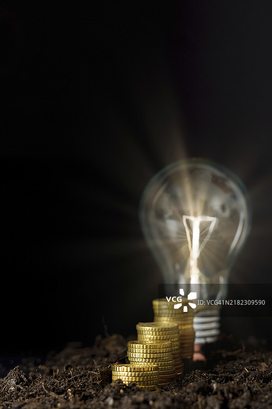 堆积如山的硬币与黑色背景上的亮灯泡，象征着电价上涨的概念图片素材