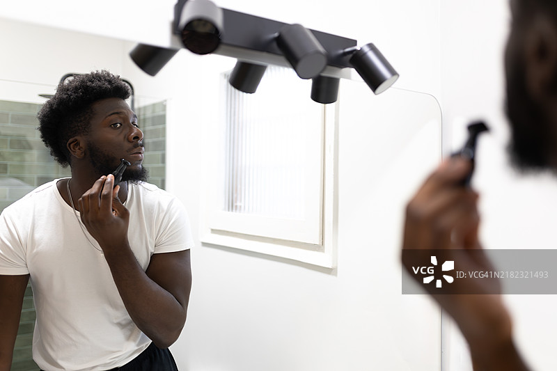 非裔美国男性在家浴室里修剪头发，镜子中映出他的身影。图片素材