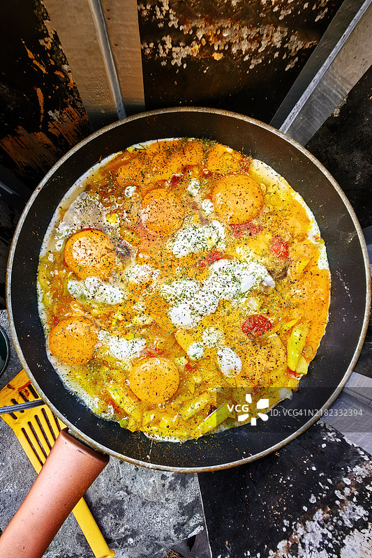 在露营炉上，鸡蛋与蔬菜和香料一起在平底锅中烹饪，周围环绕着乡村风格的户外元素和烹饪工具。图片素材