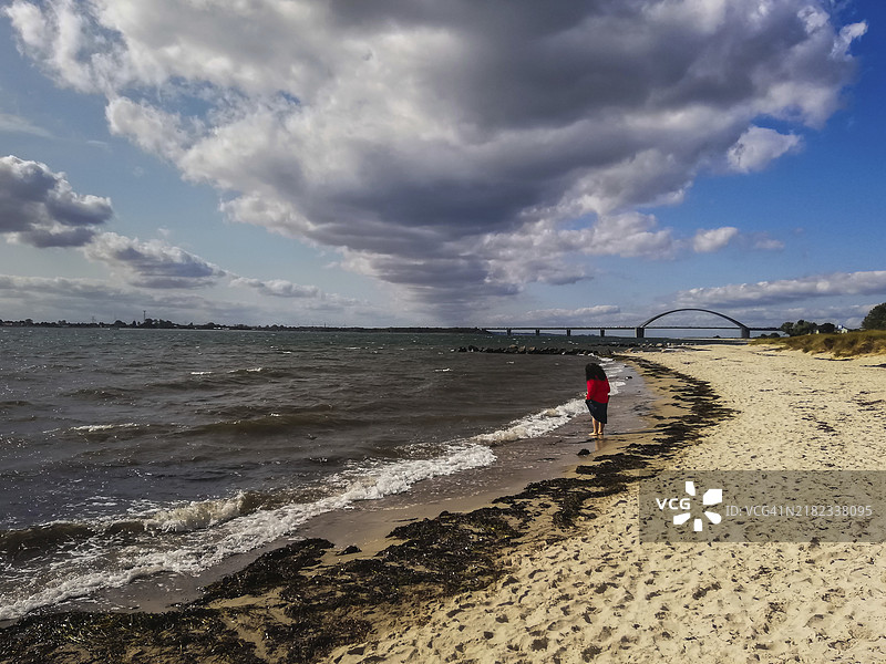 一个人在沙滩上散步，背景是桥和多云的天空，波罗的海岛屿费尔曼，费尔曼海峡，德国图片素材