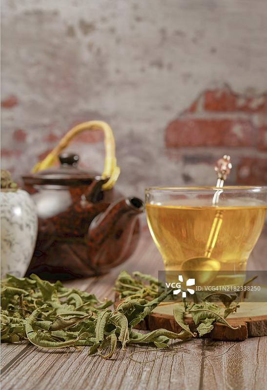玻璃杯中的柠檬香蜂草浸泡液，旁边有大理石研钵和杵，以及一个陶瓷茶壶，茶壶里放着干燥的柠檬香蜂草叶，背景是砖墙的木桌。图片素材