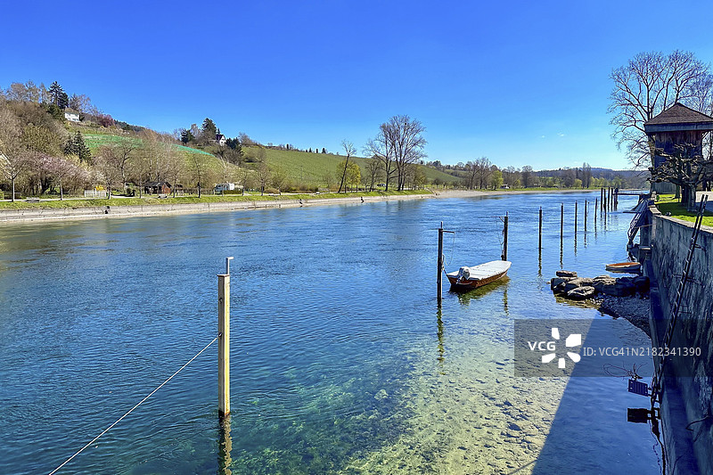 明亮阳光下的清澈河流风景，平静的水面，船只停靠处，背景是绿色的山丘，瑞士图尔高州迪森霍芬。图片素材