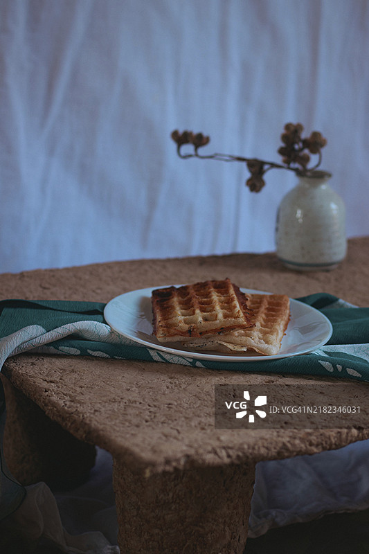 温暖的华夫饼放在白色盘子里，底下是绿色布餐巾，背景是一个装有干花的花瓶的乡村风格桌子。图片素材