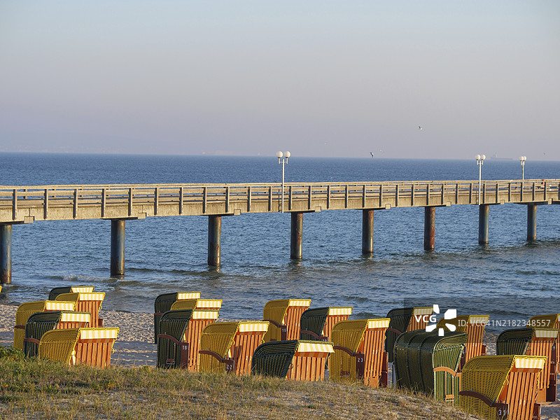 海滩上的沙滩椅,前方是一条通向大海的长码头,夕阳的温暖色调,秋季氛围,德国宾茨的海滩上,早晨的阳光照耀着沙滩椅和树木。图片素材