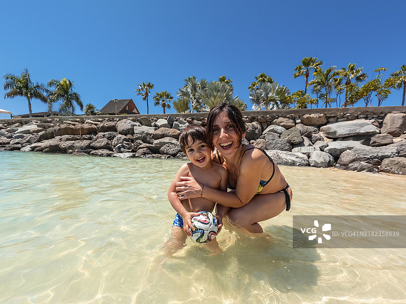 一位女性和一个孩子在水中玩耍。女性手里拿着一个球，面带微笑。场景快乐而充满玩乐气氛。图片素材