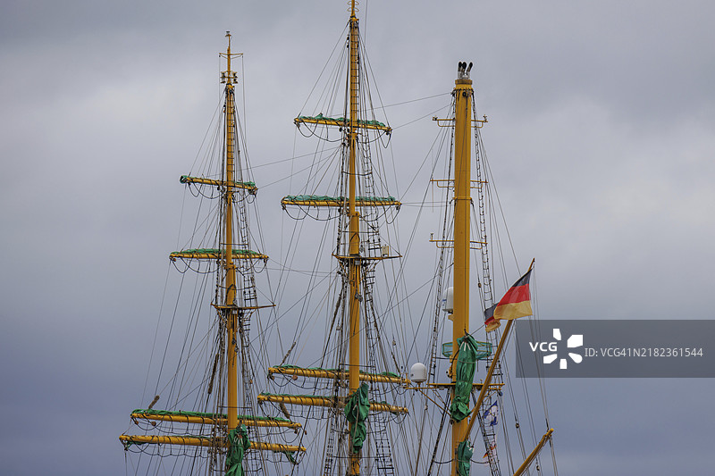 一艘帆船的桅杆上飘扬着德国国旗，背景是多云的天空，位于德国不来梅的布雷默哈芬港口。图片素材