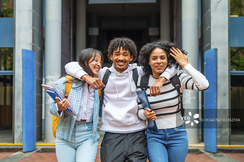 三位多民族朋友在大学里相拥在一起，面带快乐的笑容看向镜头。图片素材