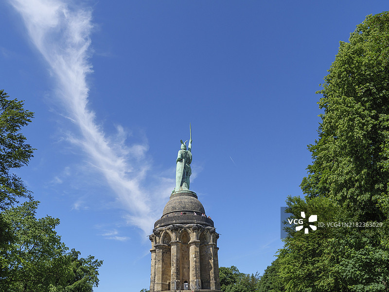 一座高大的雕像手持剑，周围环绕着树木，蓝天上飘着轻云，位于德国北莱茵-威斯特法伦州的德特莫尔德。图片素材
