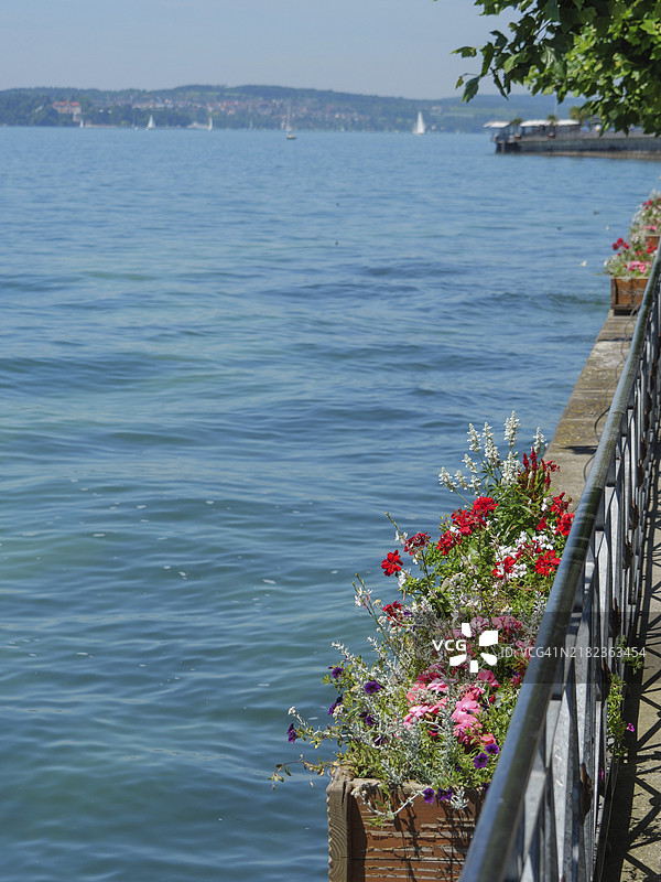 德国梅尔斯堡的湖边风景画，湖边栏杆上盛开着各种花朵，湖面上漂浮着小船，夏日的天空宁静而明朗，湖边的花箱里种满了各种花卉。图片素材