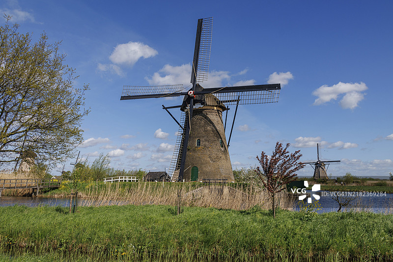一座风车雄伟地矗立在清澈蓝天和云朵下的自然泉水景观中，河边的风车，蓝天与田野上的云朵，荷兰金德代克。图片素材