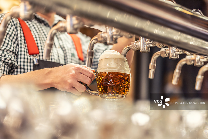 一个不明身份的人手握着一个倾斜的啤酒杯，正在倒入金色的拉格啤酒，形成泡沫和气泡。图片素材