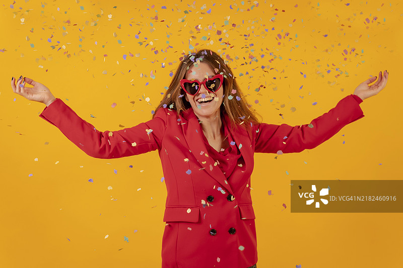 穿着红色连衣裙的女人在黄色背景下微笑，抛洒着五彩纸屑，派对概念图片素材