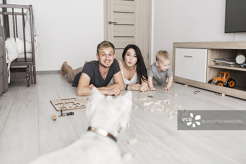 微笑的年轻夫妇躺在硬木地板上，和他们的儿子一起看着白色的狗。图片素材