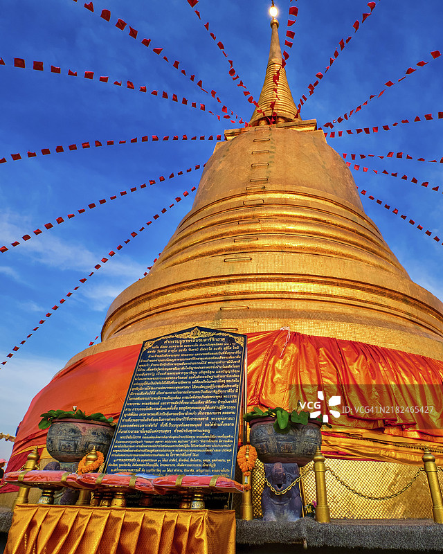 在泰国曼谷的金山寺（Wat Saket）前，一块石板上用泰语写着长长的信息，背景是装饰着节日红旗和纺织品的大金塔（chedi），夕阳的光辉洒在上面。传统的供品、祈祷、崇拜和信息。图片素材
