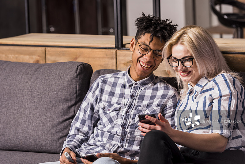 微笑的年轻情侣坐在沙发上看手机图片素材