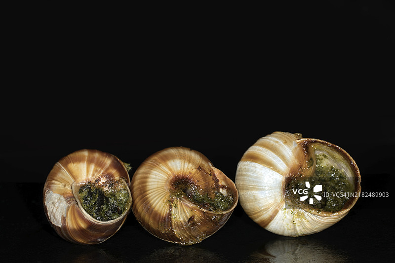 带条纹的罗马蜗牛（Helix lucorum）与香草大蒜黄油，黑色背景下的摄影作品图片素材
