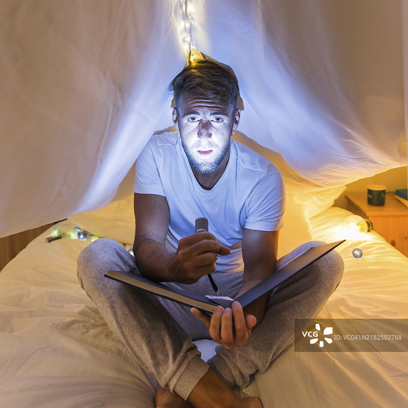 男人坐在窗帘床上，手持相册，用手电筒照亮他的脸。图片素材
