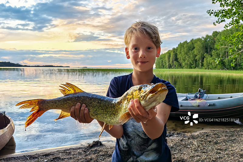 自豪的小男孩在日落时分的湖岸边举着刚捕到的梭鱼图片素材