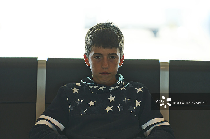 一个年轻男孩坐在机场椅子上，表情有些严肃，背着背包，等待并看向镜头，正面视角。图片素材
