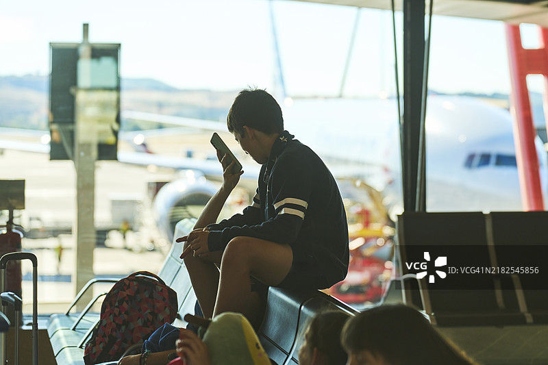 侧面视角的男孩穿着休闲服，坐在机场椅子上，和家人一起看手机。图片素材
