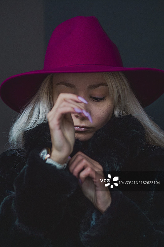 白人铂金金发女性，戴着红色帽子，沉思地低头，灯光氛围独特。图片素材