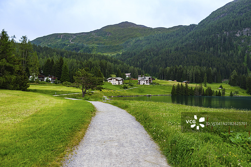 绿色草地上有小路，四周环绕着山脉和几座房屋，天空多云，瑞士达沃斯，欧洲图片素材