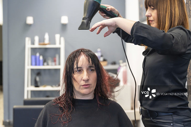 一位严肃而耐心的女性坐在发廊里，发型师正在为她吹干头发。图片素材