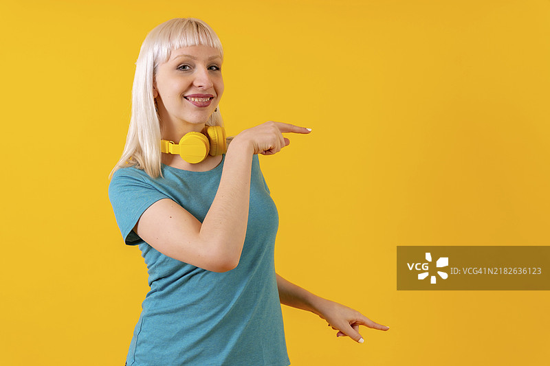 戴着耳机的金发白人女孩在黄色背景的摄影棚中指向右侧的广告图片素材