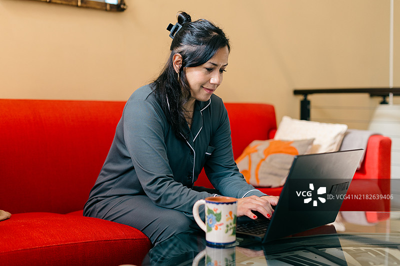 一位混血的西班牙裔拉丁裔亚裔女性穿着睡衣在家中用笔记本电脑工作，旁边放着一杯咖啡。这是一个在家工作的日子，或者是一位母亲在一天开始前处理电脑任务。图片素材