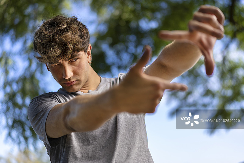 一位英俊的年轻男子眨眼，透过由手组成的框架看去。迷人的男子用手指做成框架，背景模糊，留有文本空白。图片素材