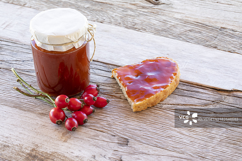 自制玫瑰果酱装在罐子里，旁边是一片涂抹了果酱的面包，放在木桌上，周围有成熟的水果。图片素材