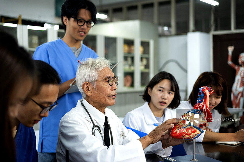 一位经验丰富的年长亚洲男性教授在解剖教室中使用人体和器官模型教授人体解剖学，向年轻的护理和医学学生分享他的专业知识和经验。图片素材