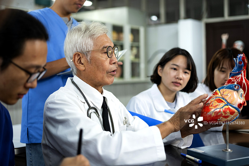 一位经验丰富的年长亚洲男性教授在解剖教室中使用人体和器官模型教授人体解剖学，向年轻的护理和医学学生分享他的专业知识和经验。图片素材