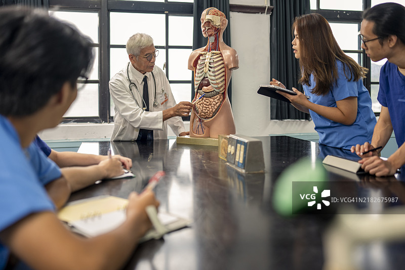 一位经验丰富的年长亚洲男性教授在解剖教室里使用人体和器官模型教授人体解剖学，向年轻的护理和医学学生分享他的专业知识和经验。图片素材