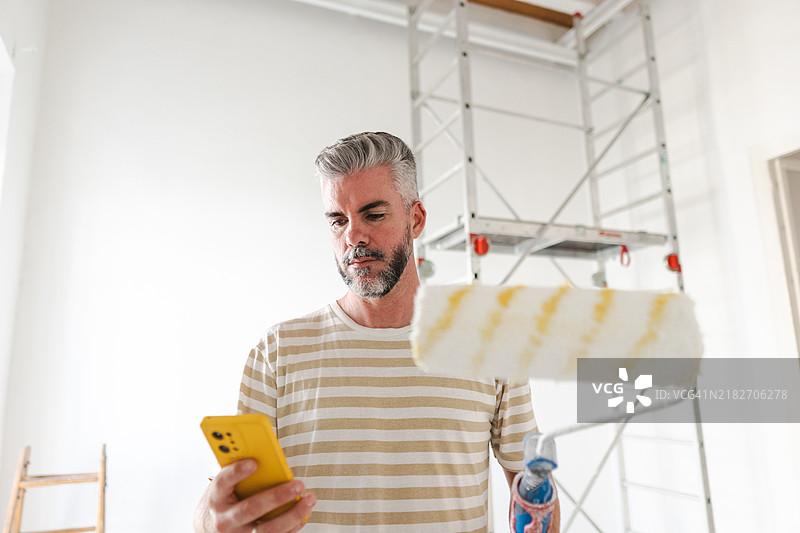 在翻新房间中，画家一手拿着智能手机，一手握着油漆滚筒。图片素材