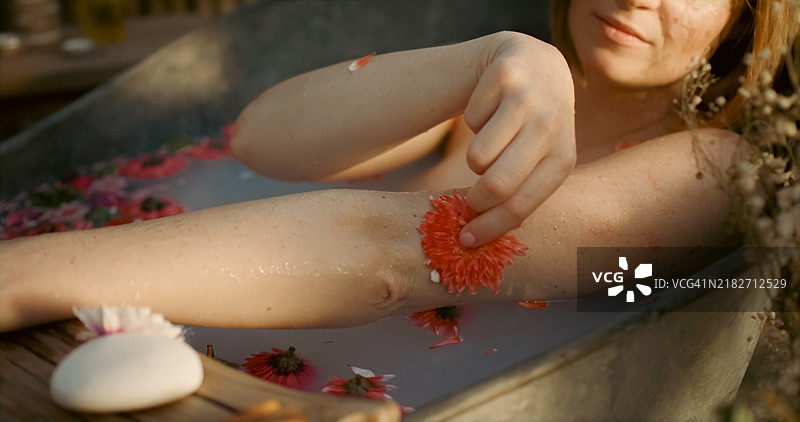 一位红发女性用花朵轻抚自己的手。她坐在一个锌制浴缸里。图片素材