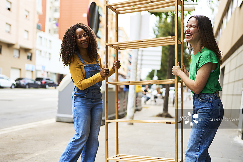 两位女性在街上侧身站立，边谈边笑，旁边有一个用于再利用的木架子。图片素材