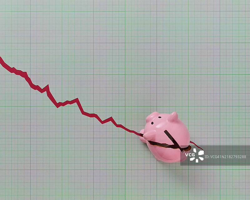 一只粉色的储蓄罐放在方格纸上，上面有一道撕裂，像是向下的财务图表。图片素材