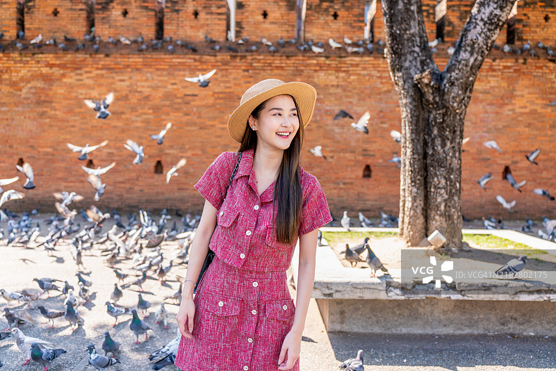 在清迈塔佩门的历史砖墙旁，一位戴帽子的亚洲女性与飞翔的鸽子享受着美好时光。图片素材