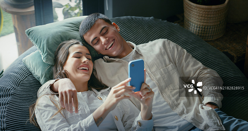 拥抱、欢笑的情侣与智能手机，在家中通过社交媒体分享视频，温馨的约会。男人和女人使用手机，快乐地观看订阅的电影，搞笑的家庭时光。图片素材