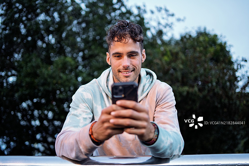 穿着休闲扎染连帽衫的快乐年轻男子在户外使用智能手机并微笑图片素材