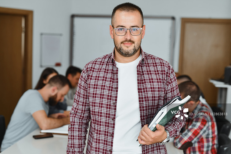 男教授微笑着手持书籍，面向镜头，背景中有学生在写作。图片素材